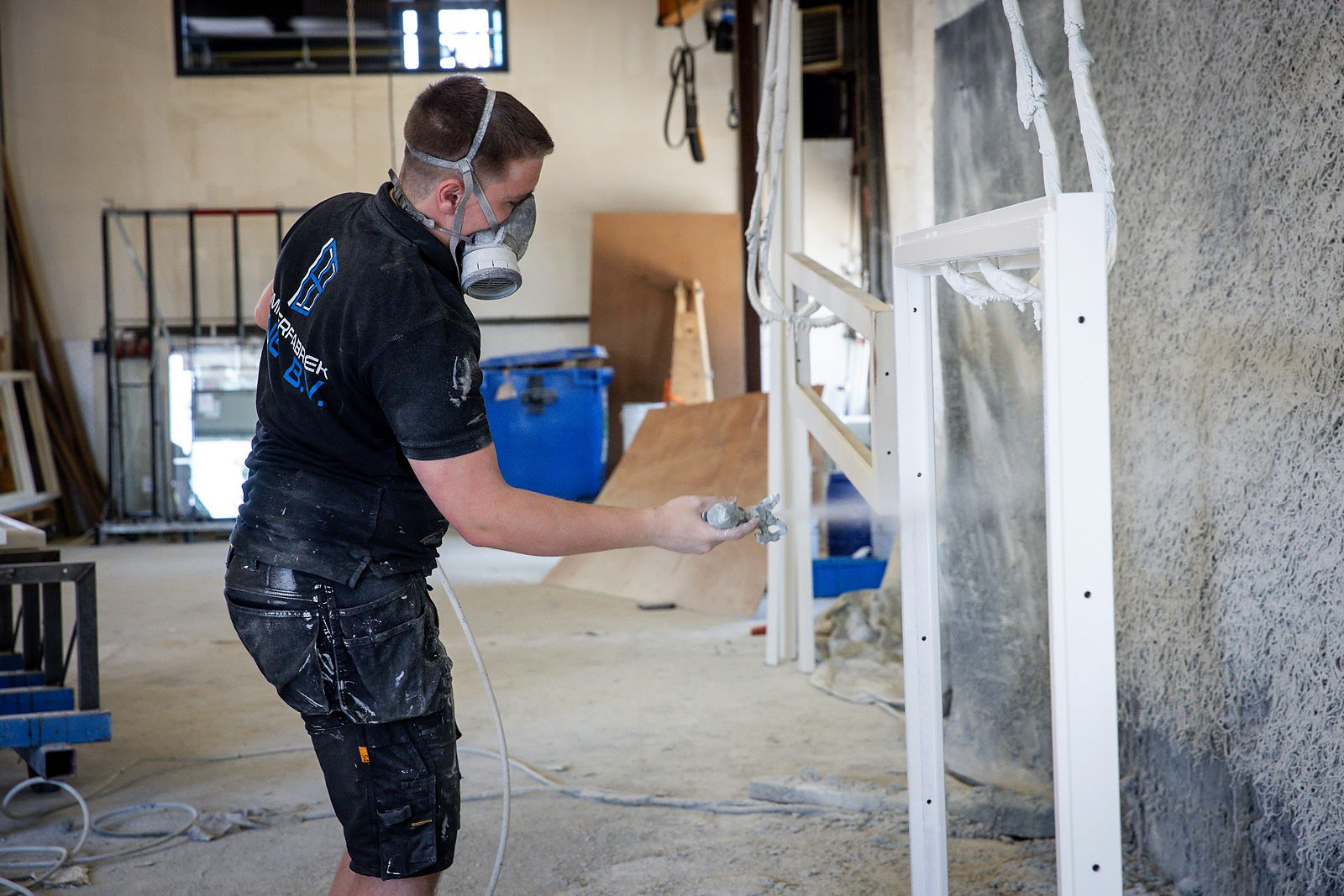 Medewerker met ademmasker spuit in de spuiterij van de werkplaats een wit kozijn met een spuitpistool; rechts een spuitwand vol opgedroogde verf.