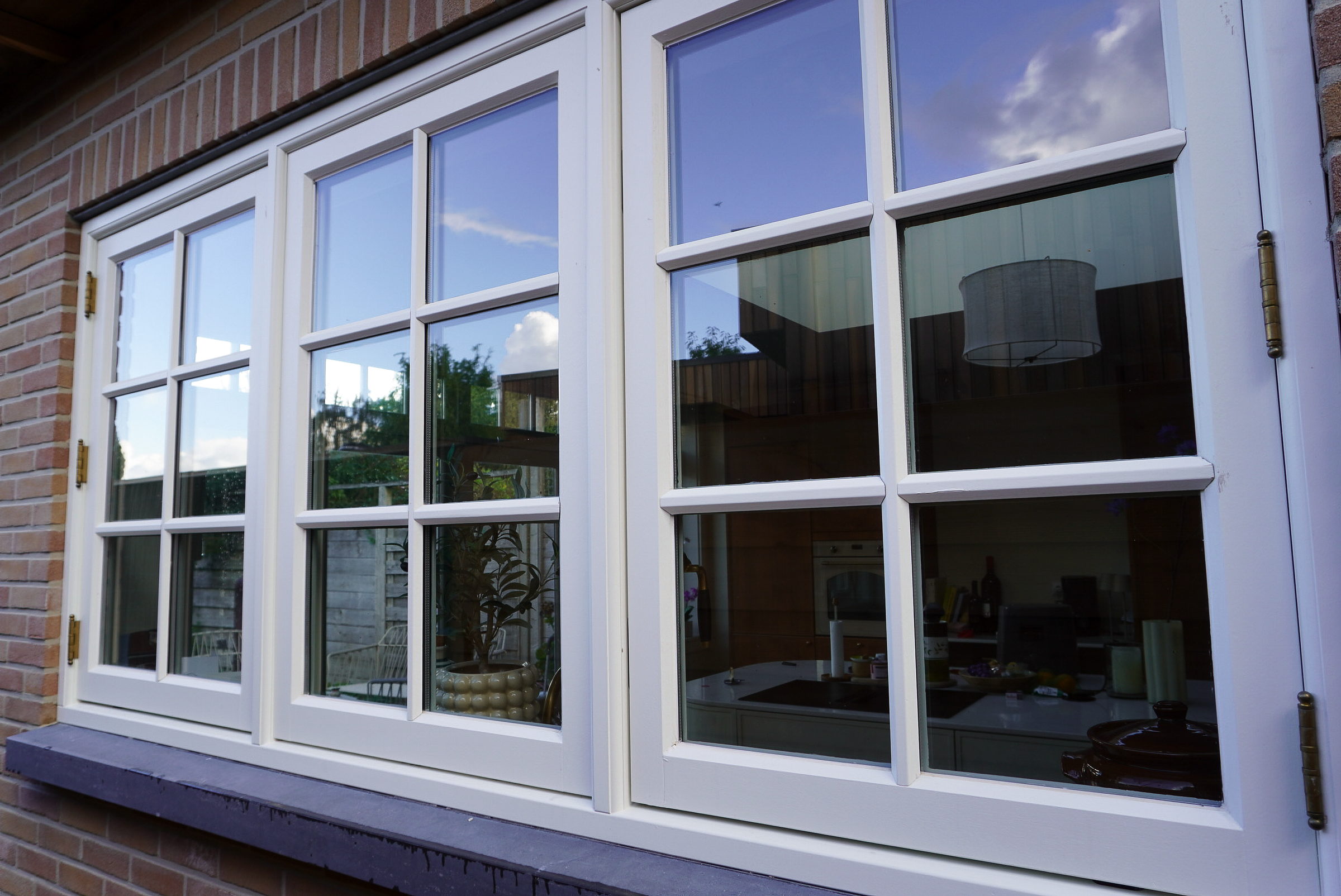 Driedelig wit houten kozijn met roedeverdeling en messing scharnieren, gevat in een bakstenen gevel op een donkere natuurstenen onderdorpel; in het glas spiegelen lucht en bomen.